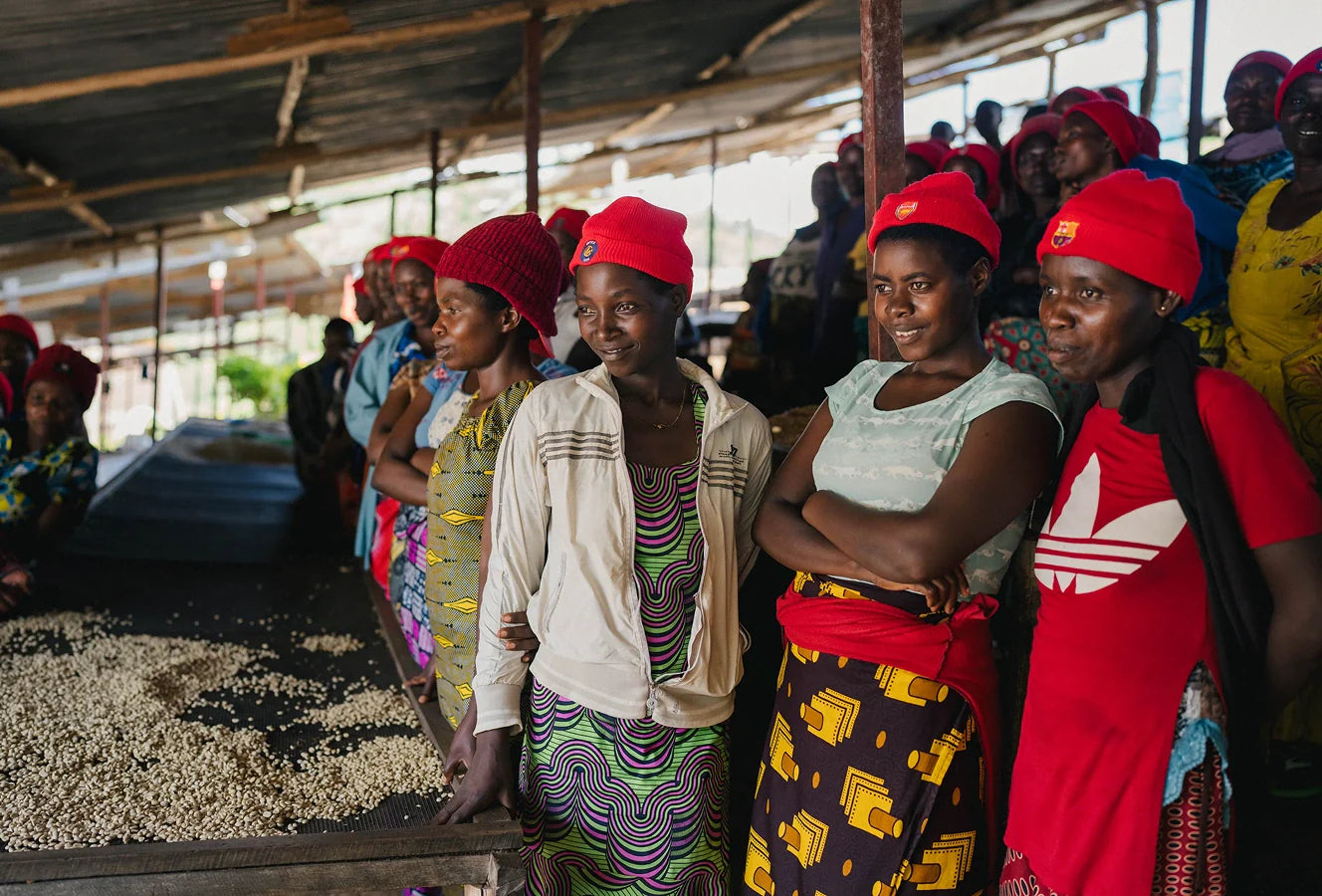 A group of women wearing red hats, as part of the Rwanda Sholi Kundwa Women Coffee Collective, at a coffee process setting.
