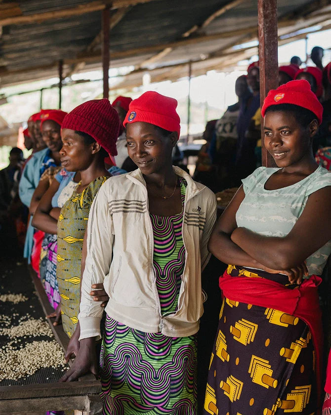 A group of women wearing red hats, as part of the Rwanda Sholi Kundwa Women Coffee Collective, at a coffee process setting.