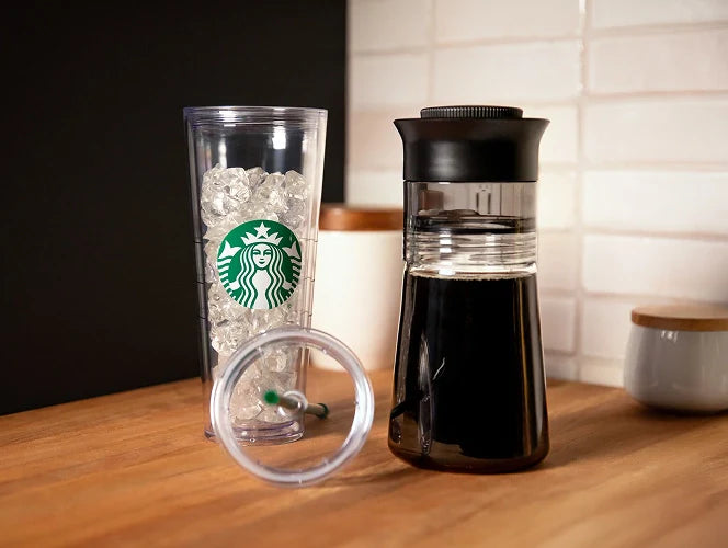 A Starbucks Plastic Cold Cup filled with ice, and a Cold Brewer filled with coffee placed on a wooden tabletop, likely in a kitchen setting.