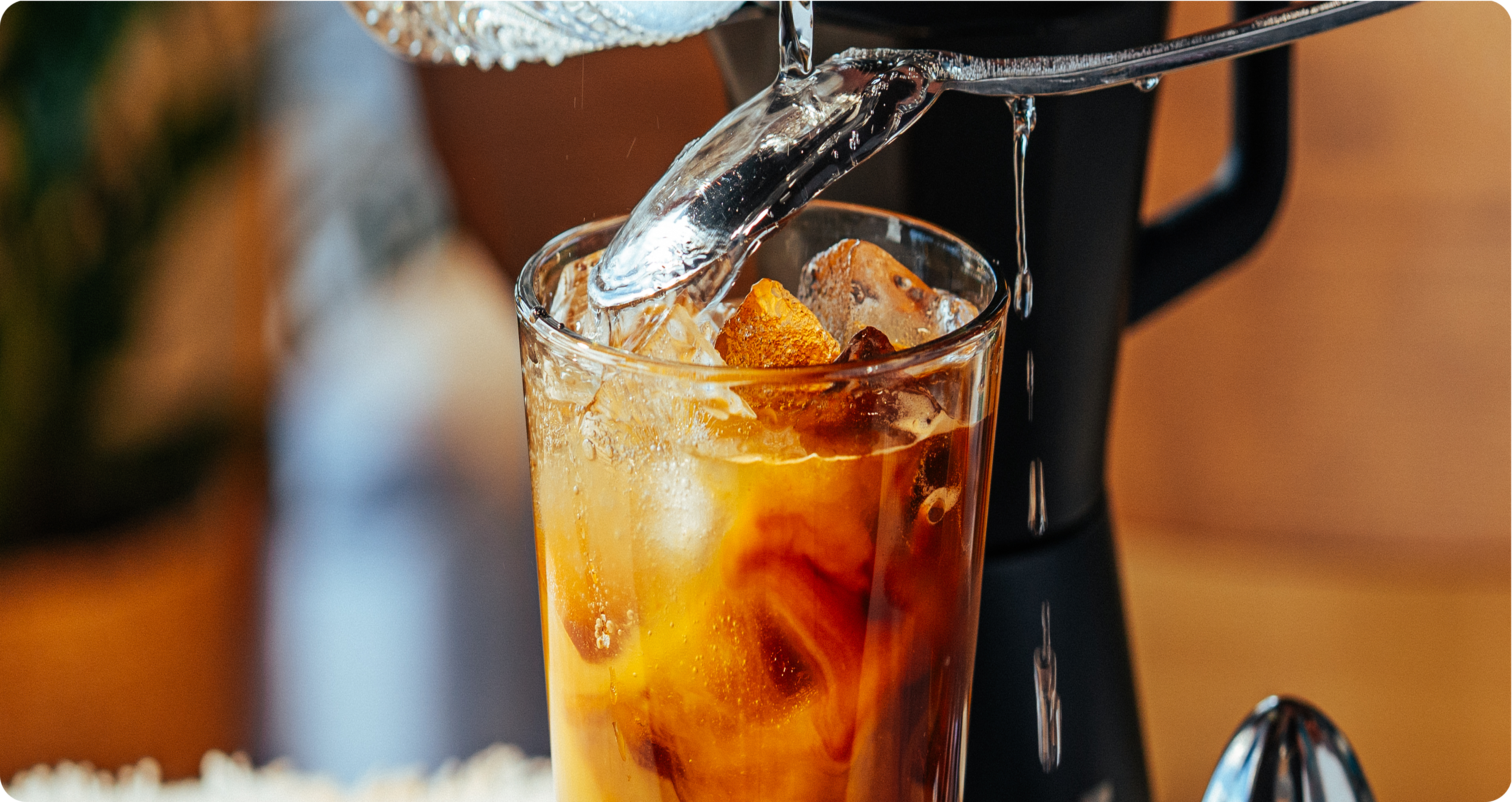 A close-up of iced coffee in a clear glass with ice cubes, while hot water is being poured from a metal pitcher over the top. The background is softly blurred with warm indoor tones.