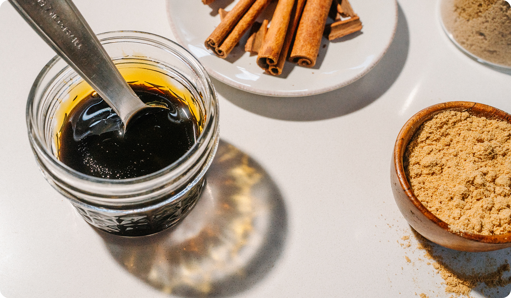 A close-up of a clear glass jar filled with dark brown syrup or honey, with a metal spoon resting inside. The jar sits on a white surface with cinnamon sticks on a small white plate and a wooden bowl of light brown powder nearby.