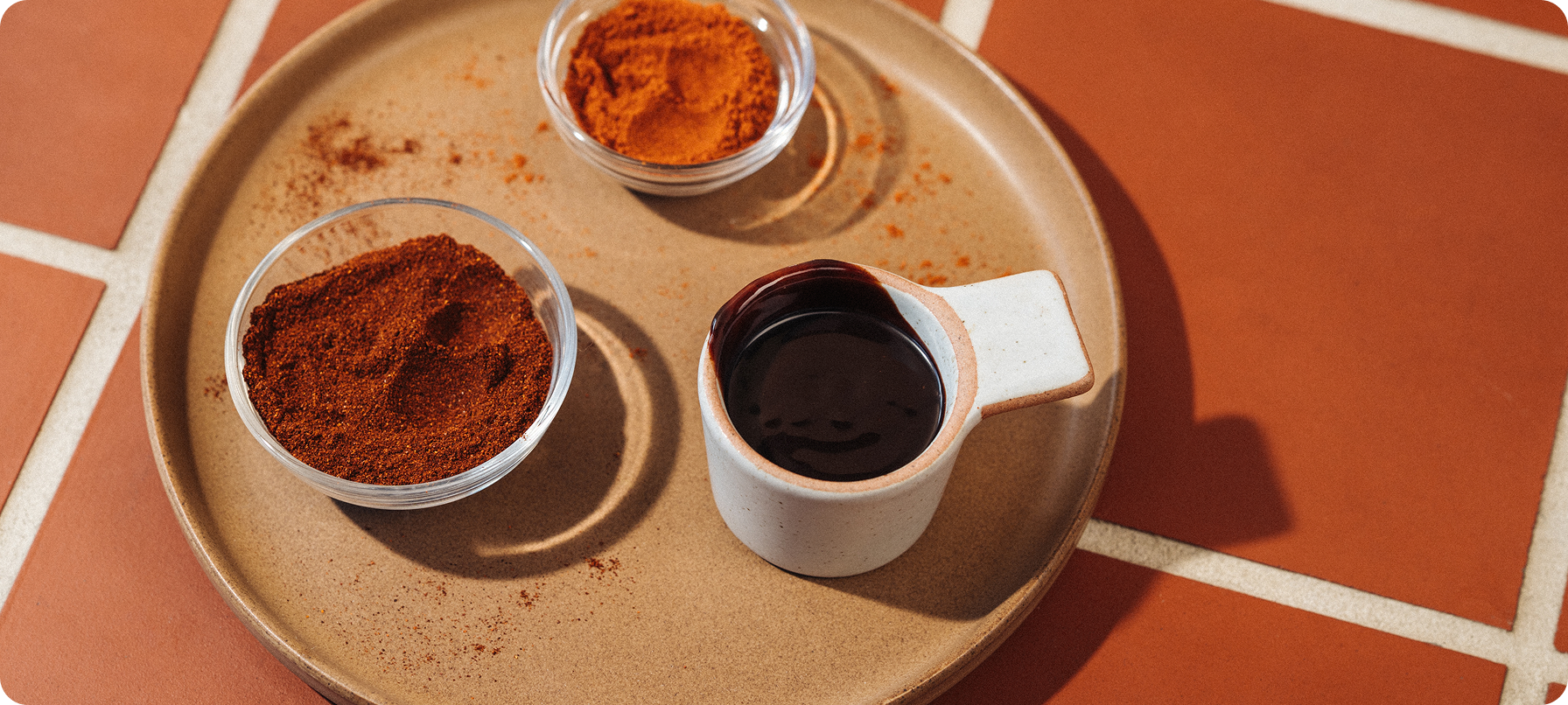 A beige ceramic tray on terracotta tile, holding three small containers: two clear bowls with reddish-brown powders and one small white cup with a dark brown liquid.