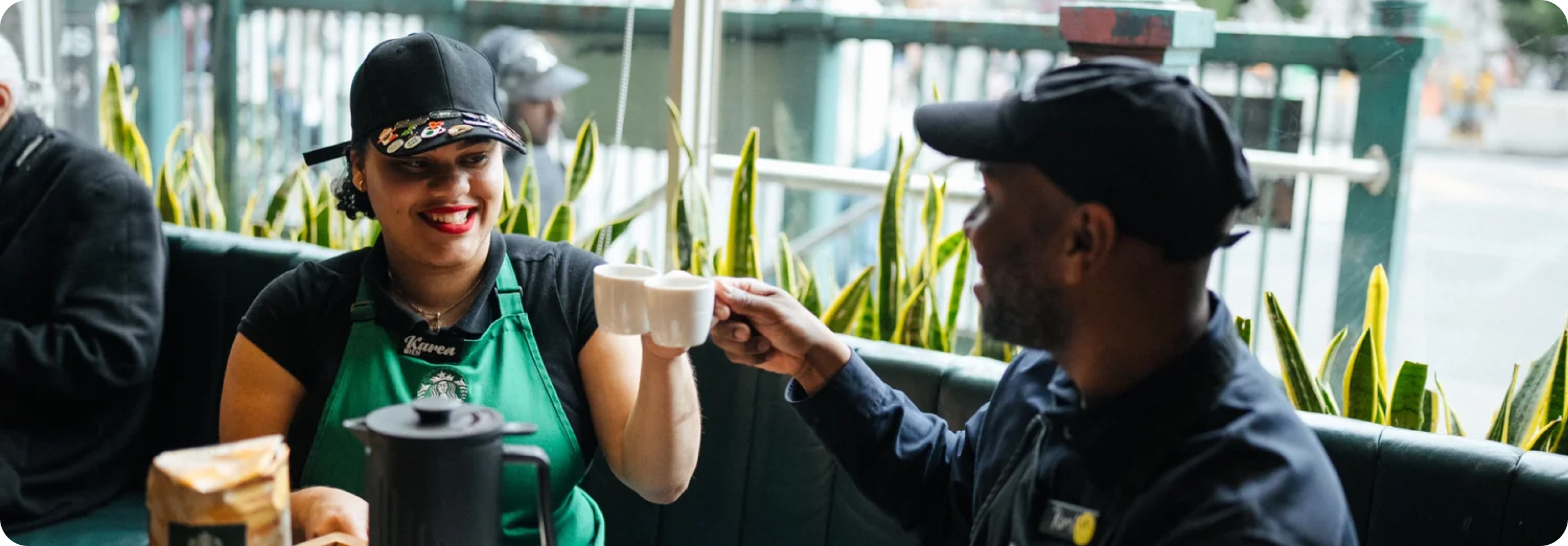 Person in a green apron interacting with another person outdoors, with plants and a blurred background.