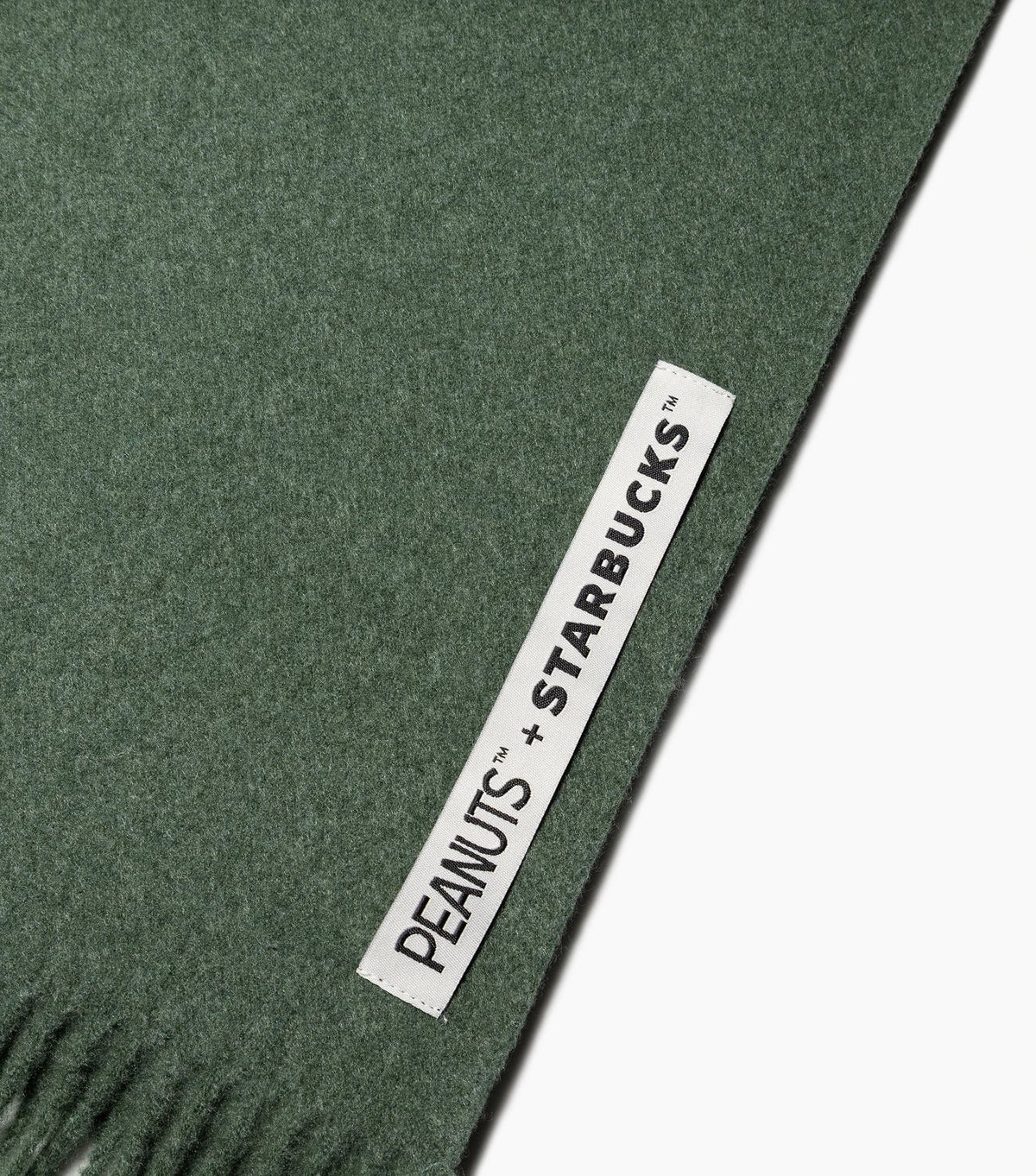 A close-up of a dark green woven textile with a white fabric label reading 'PEANUTS + STARBUCKS' printed in black, shown on a white background.