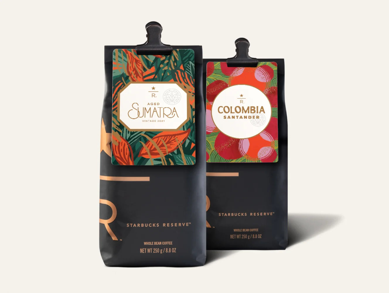 Two Starbucks Reserve coffee bags standing upright on a light beige background. The left bag is labeled aged Sumatra with a green tropical leaf pattern, and the right bag is labeled Colombia Santamaria with a red strawberry pattern.