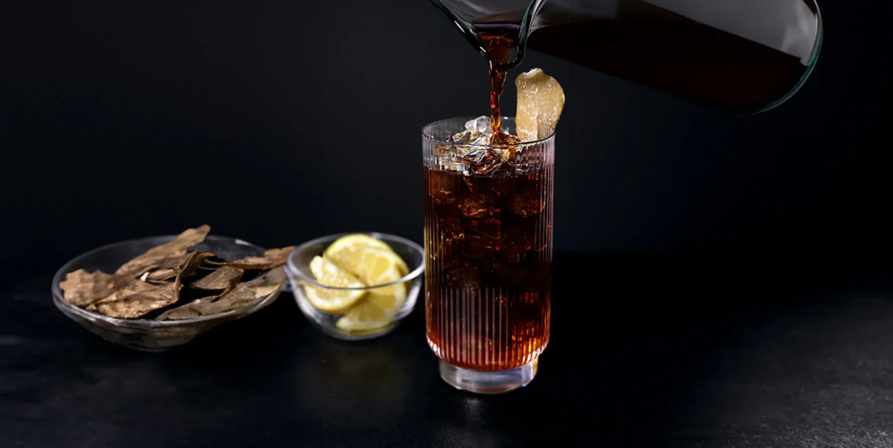 Coffee being poured into a glass of ice garnished with candied ginger, with dried lemon peel and lemon in the background.