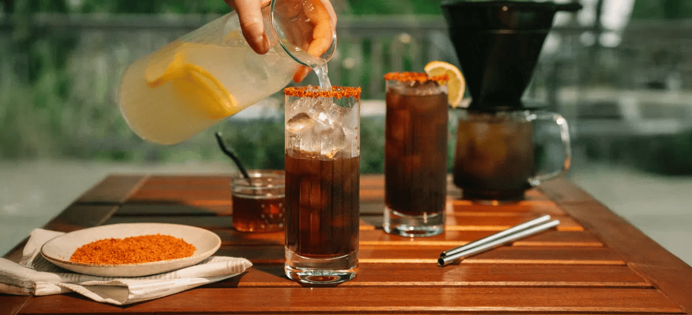 Person pouring a Hot Honey Coffee Lemonade into a glass on a wooden table with outdoor setting.