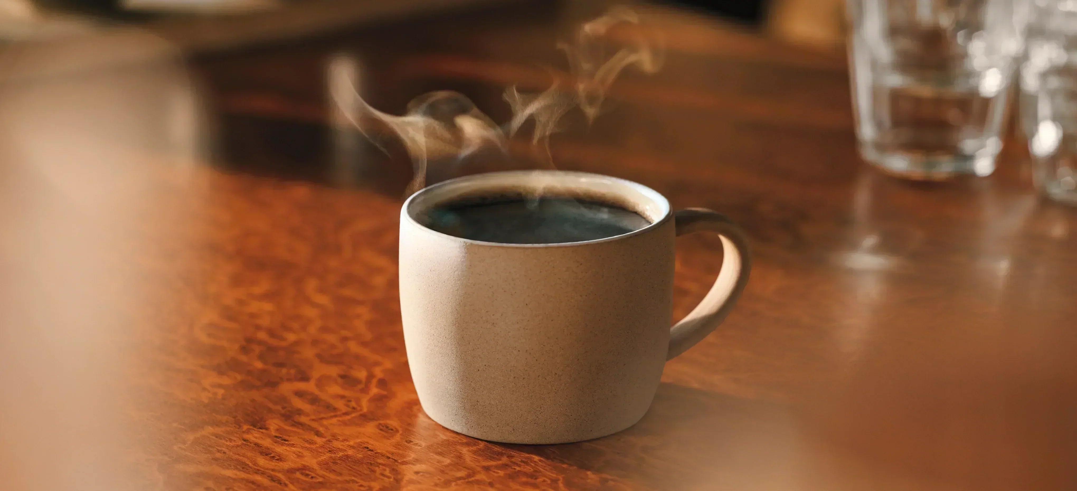 A steaming cup of black coffee in a stoneware mug on a polished wooden table.