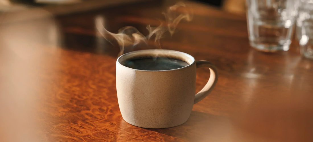 A steaming cup of black coffee in a stoneware mug on a polished wooden table.
