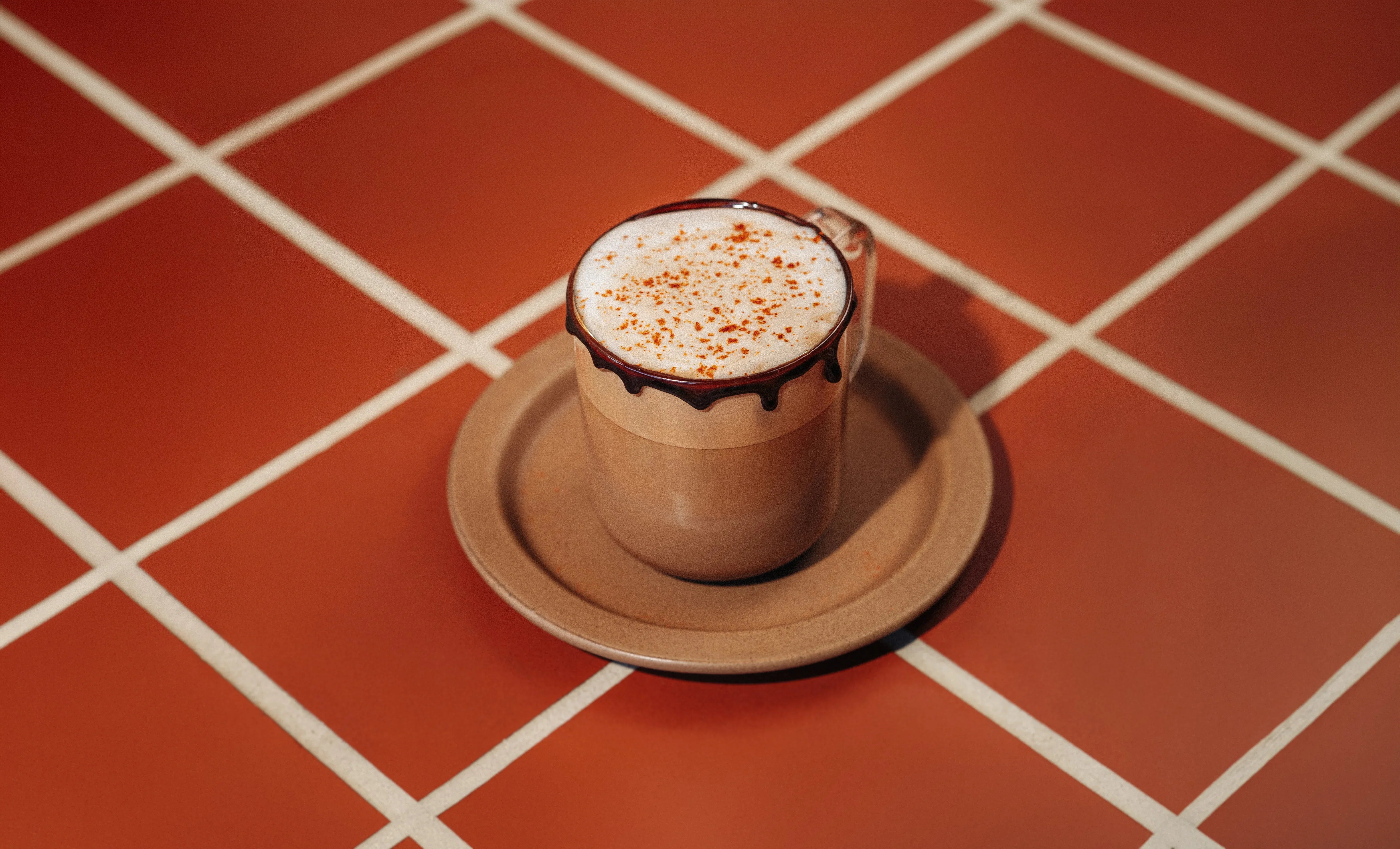 A glass mug of mocha latte with milk foam, a chocolate-dipped rim and a sprinkle of chili powder, on a saucer on red tiles.