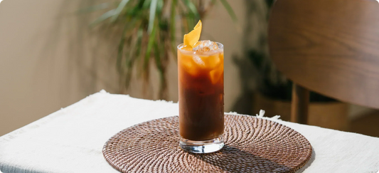 Iced sparkling orange americano drink in a tall glass with a lemon peel, on a natural woven placemat.