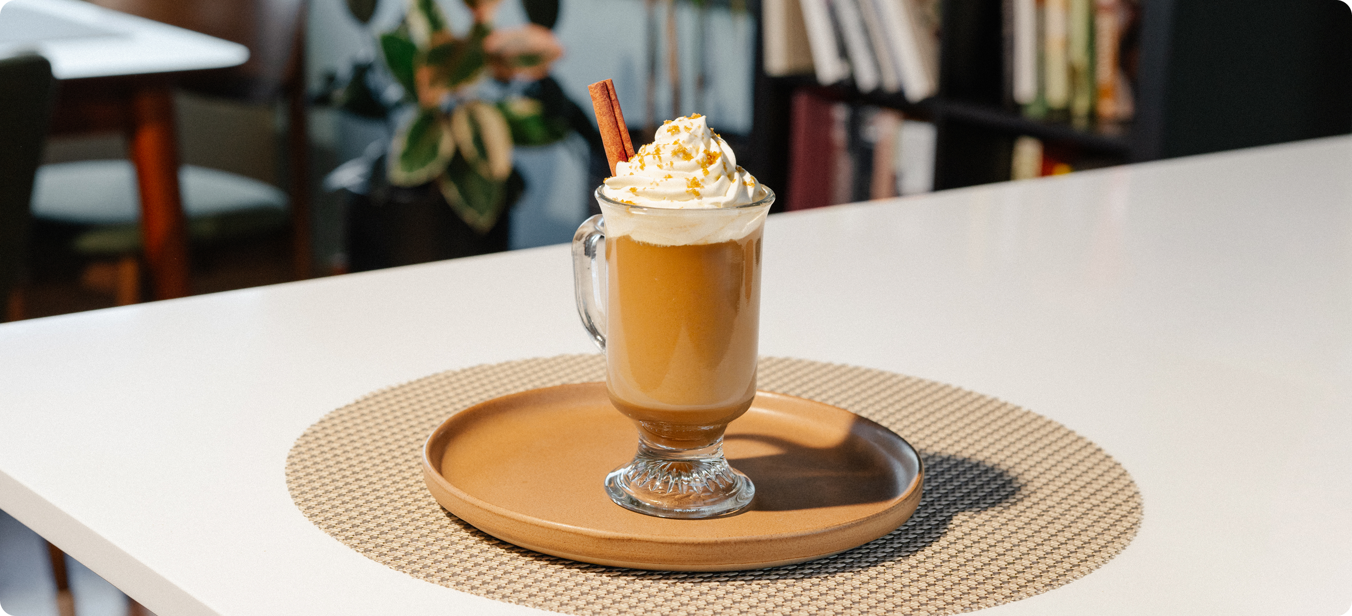 A glass mug filled with a hot molasses spice coffee drink topped with whipped cream and a cinnamon stick, set on a round wooden tray over a woven placemat on a white table in a softly blurred indoor setting.
