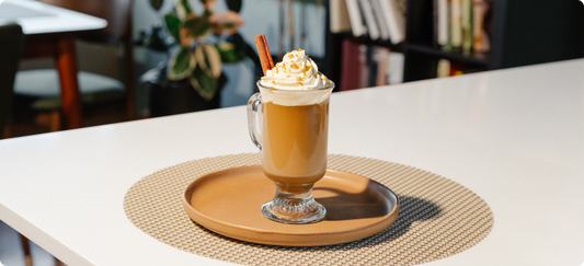 A glass mug filled with a hot molasses spice coffee drink topped with whipped cream and a cinnamon stick, set on a round wooden tray over a woven placemat on a white table in a softly blurred indoor setting.