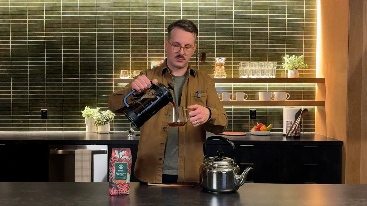 A person pours coffee from a French press into a glass at a kitchen counter, next to a kettle and a bag of Starbucks coffee.