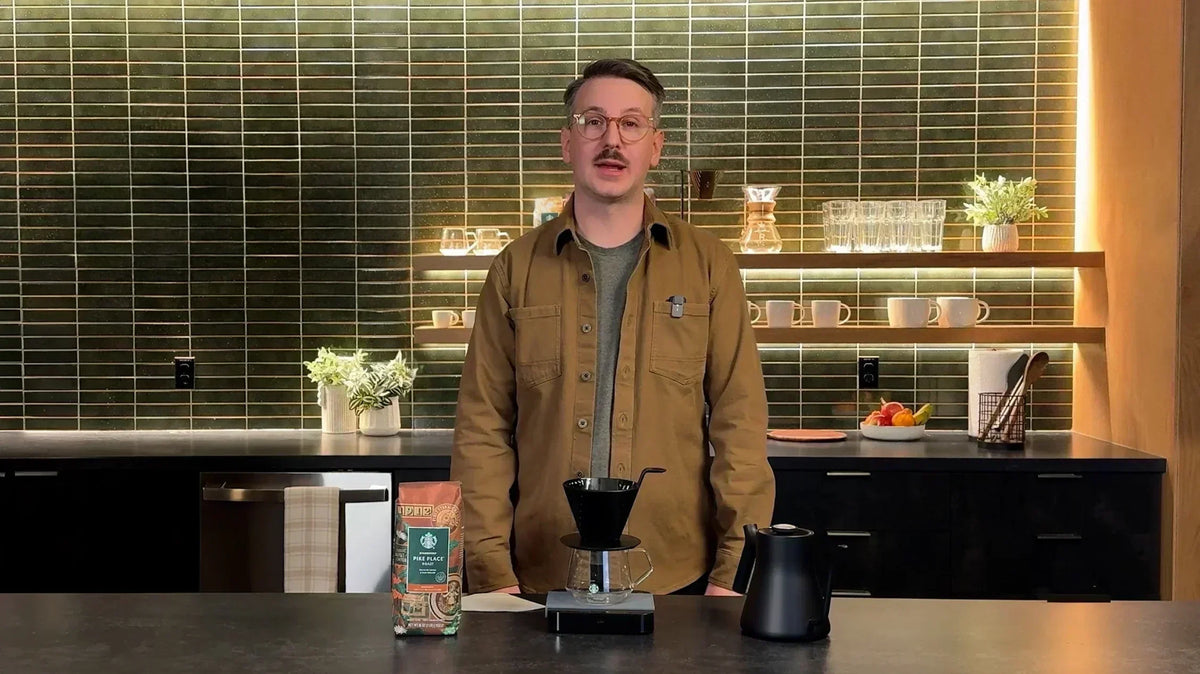 A person stands at a kitchen counter with a pour-over coffee cone on a stand, a kettle and a bag of Starbucks coffee.
