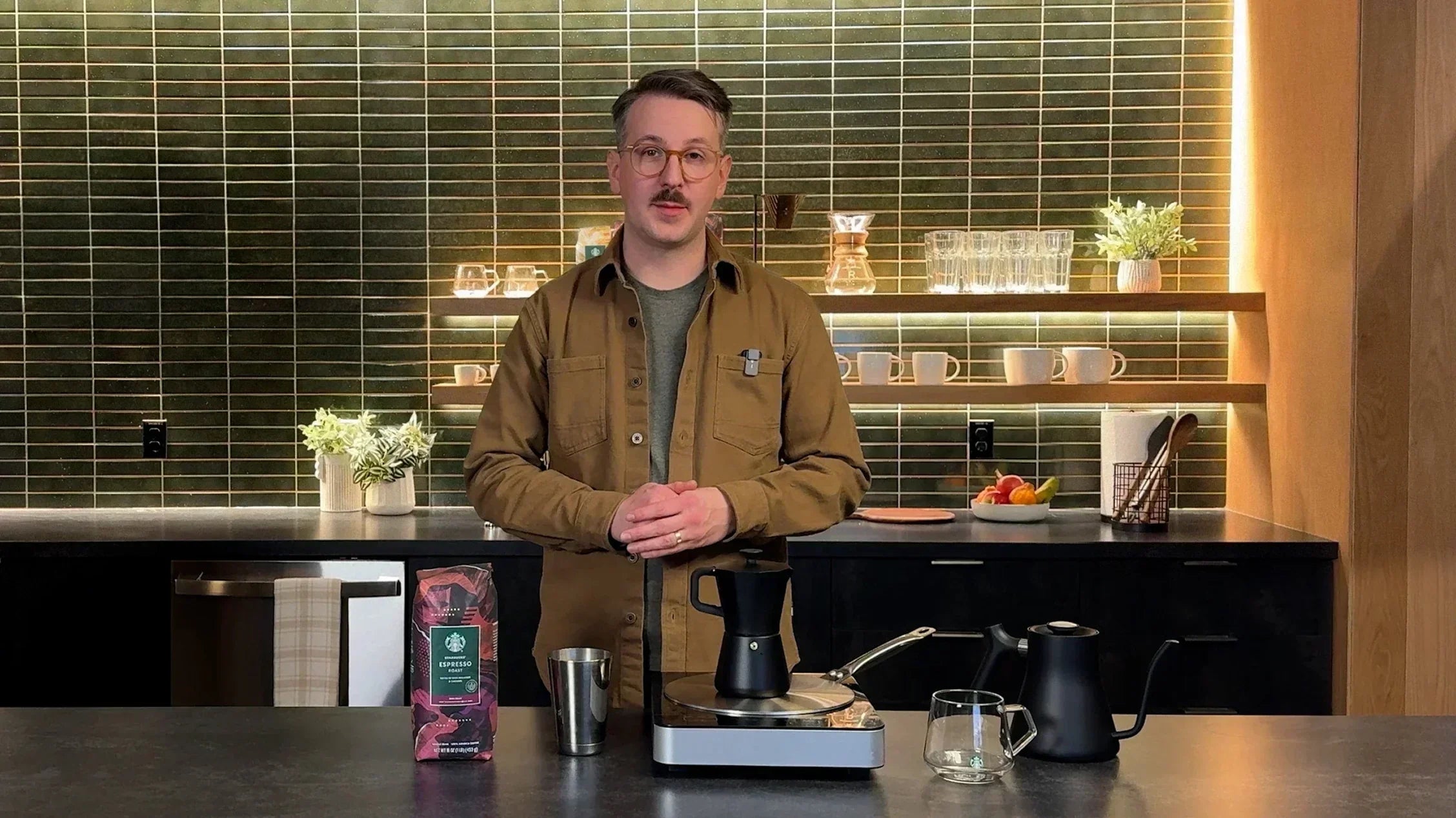 A person stands at a kitchen counter with a moka pot, a kettle and a bag of Starbucks espresso coffee.