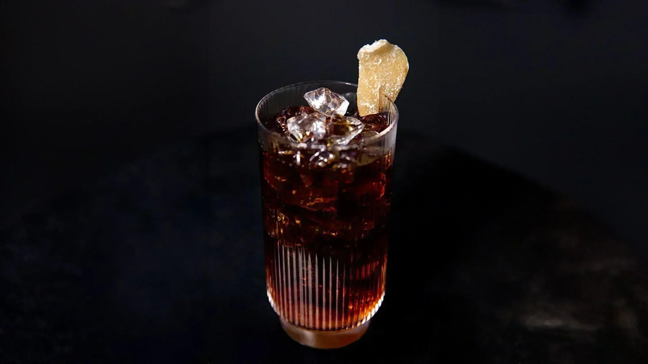 A tall glass of iced coffee over ice, garnished with a piece of candied ginger, on a dark surface.