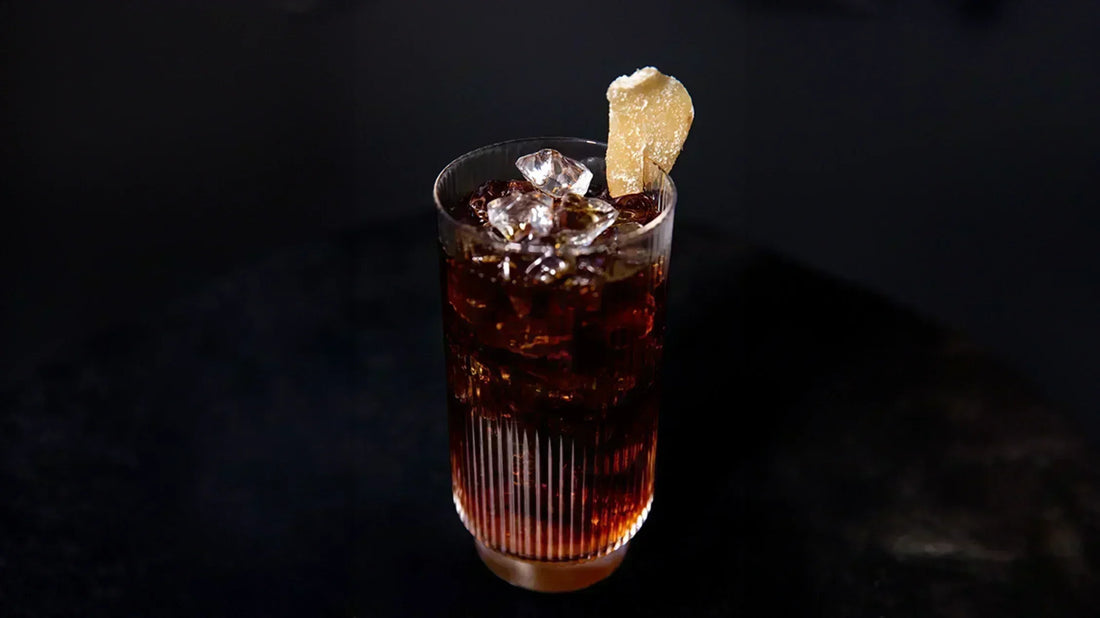 A tall glass of iced coffee over ice, garnished with a piece of candied ginger, on a dark surface.