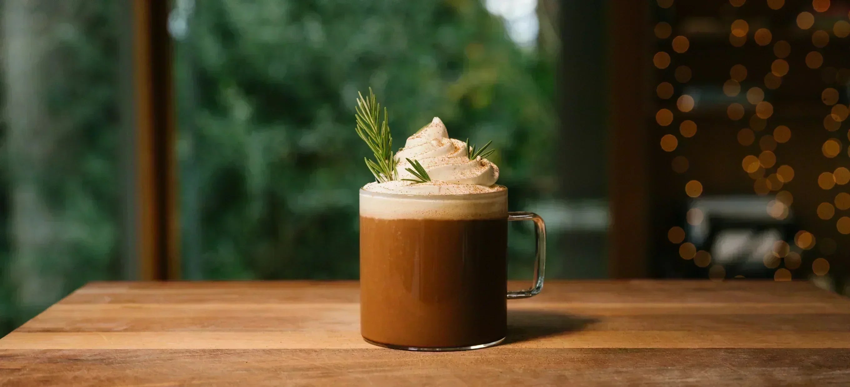 A mug of coffee topped with whipped cream, a dusting of spice and a sprig of rosemary, on a wooden table.