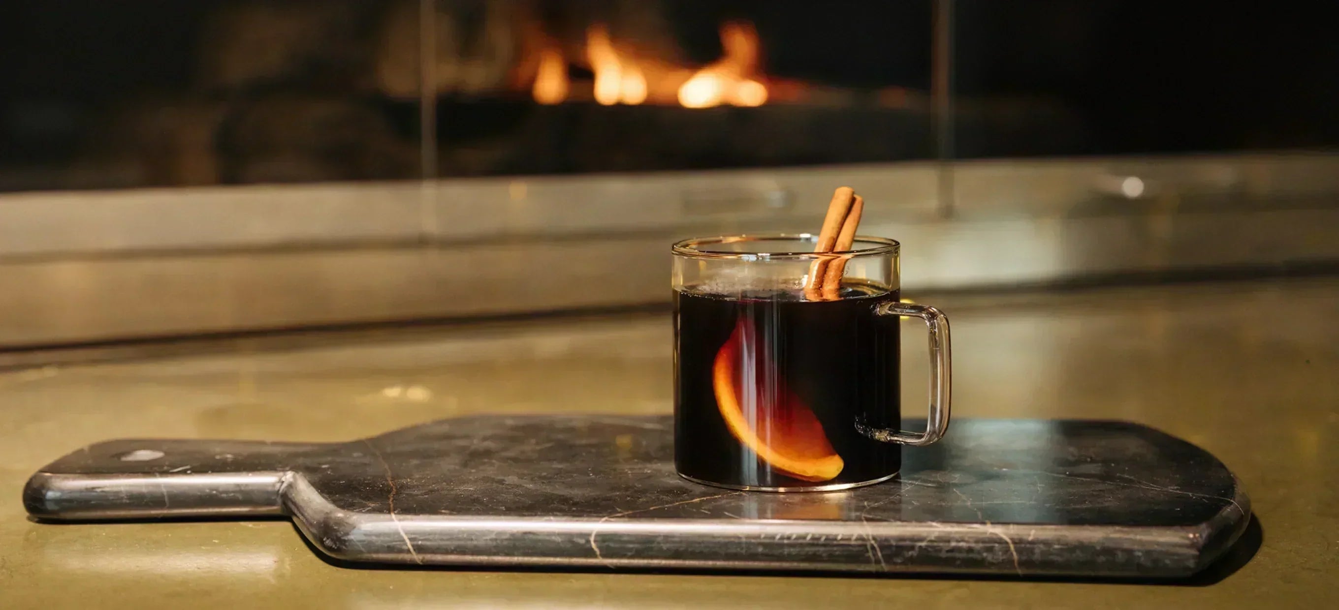 A clear mug of dark spiced coffee with an orange slice and a cinnamon stick, on a board in front of a fire.