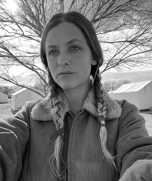 Black and white selfie photo of the artist, Becca Fuhrman with braided hair, standing in front of a tree with multiple tents in the background.