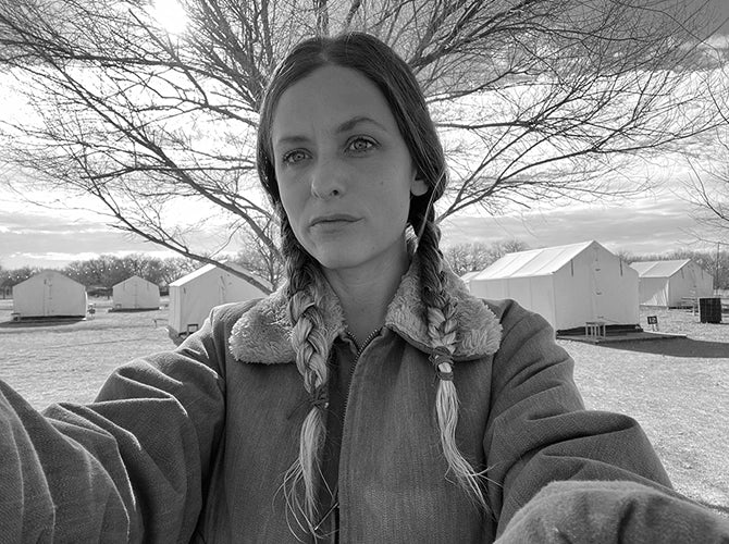 Black and white selfie photo of the artist, Becca Fuhrman with braided hair, standing in front of a tree with multiple tents in the background.