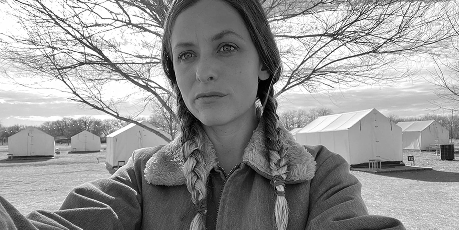 Black and white selfie photo of the artist, Becca Fuhrman with braided hair, standing in front of a tree with multiple tents in the background.