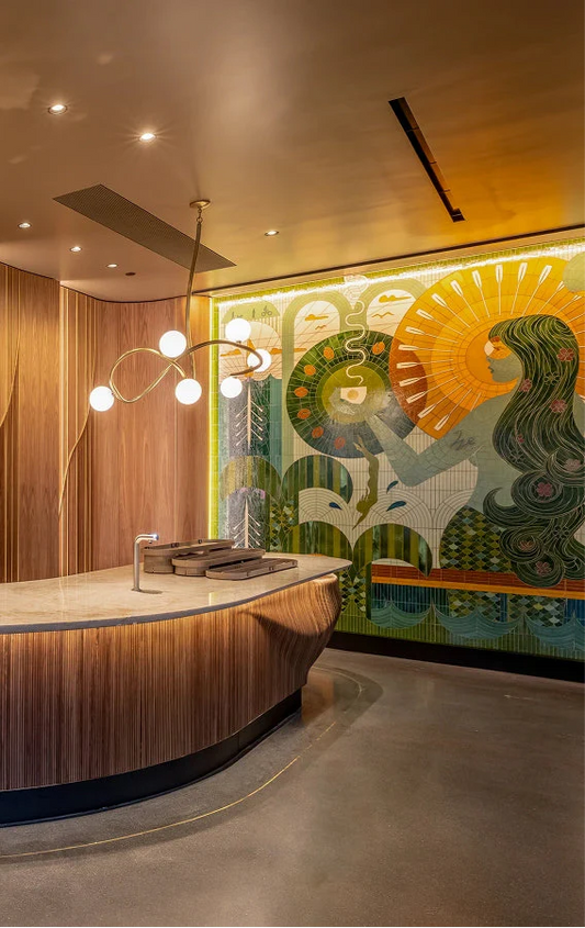A cafe counter with a curved wooden base in front of a large tiled mural of a woman with long green hair and a golden sun.