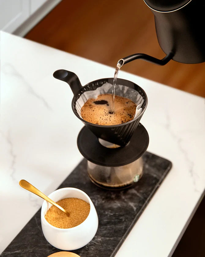 Water being poured into a filter basket, with coffee brewing through a Starbucks Plastic Pour Over, on a kitchen counter with a sugar container next to it.