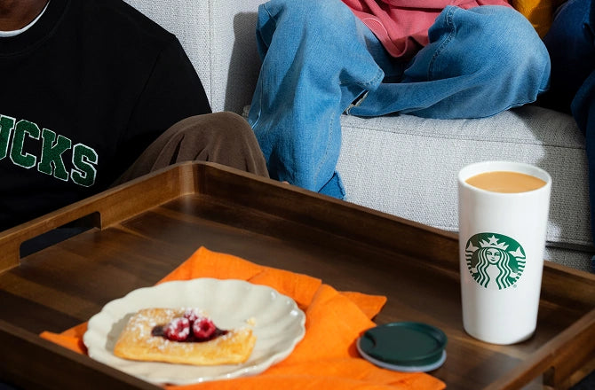 Starbucks cup on a tray with food, person sitting on a couch in the background