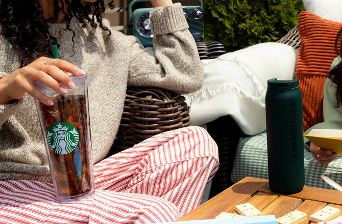 Person holding a Starbucks cup outdoors with another cup on a table.