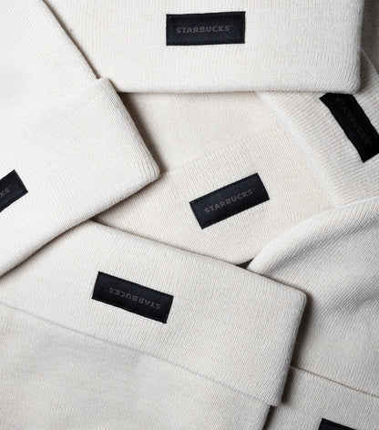 A stack of natural beige Starbucks Knit Beanies with a black Starbucks tag at the front.