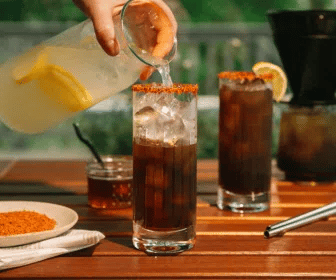 Person pouring a Hot Honey Coffee Lemonade into a glass on a wooden table with outdoor setting.