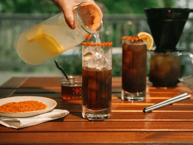 Person pouring a Hot Honey Coffee Lemonade into a glass on a wooden table with outdoor setting.