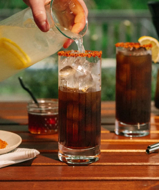 Person pouring a Hot Honey Coffee Lemonade into a glass on a wooden table with outdoor setting.