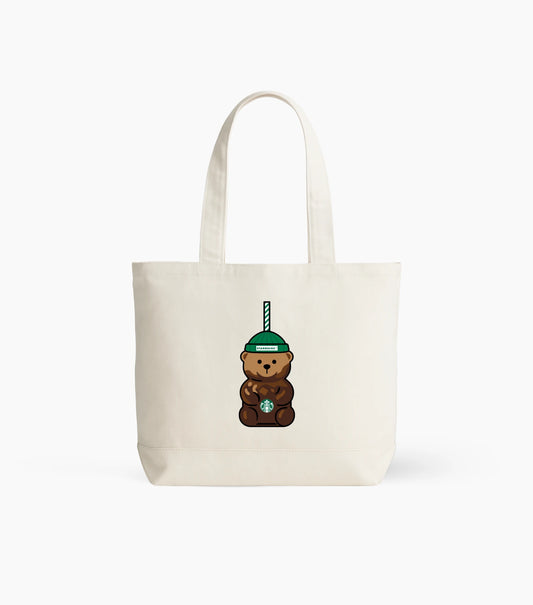 An off-white canvas tote bag with a cartoon graphic of a brown teddy bear wearing a green beanie and holding a Starbucks cup, set against a plain white background.