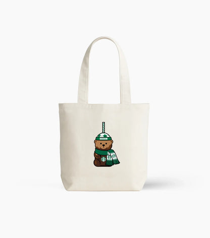An off-white canvas tote bag with a cartoon graphic of a brown teddy bear wearing a green beanie and holding a Starbucks cup, set against a plain white background.
