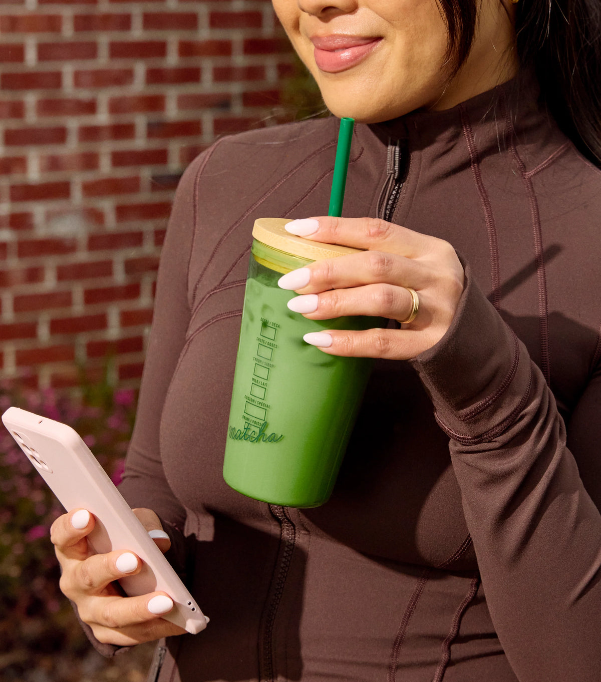 Person holding a green reusable cup with a straw and a phone, standing against a brick wall.