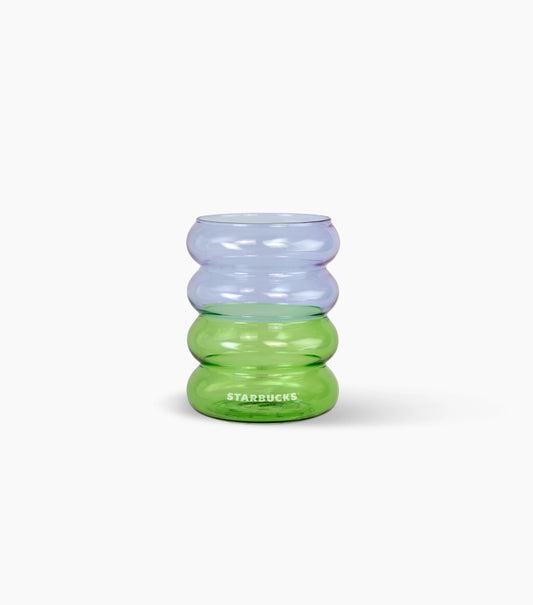 A Starbucks glass rippled tumbler with a stacked, wavy ribbed design, featuring a translucent light purple top section and a translucent green bottom section with the white Starbucks logo printed on the front, set against a plain white background.