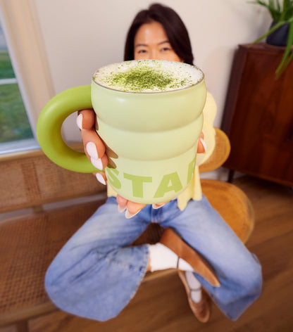 Person holding a green mug with a drink, sitting on a chair indoors.