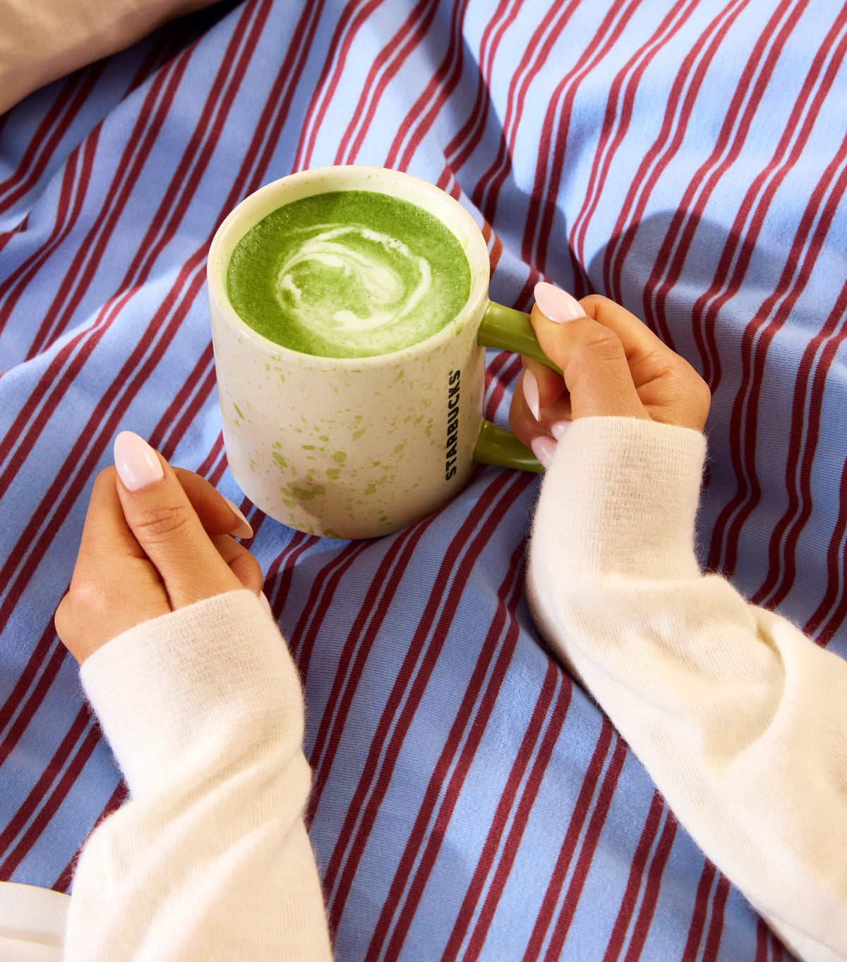 Person holding a Starbucks mug with green beverage on a striped fabric background