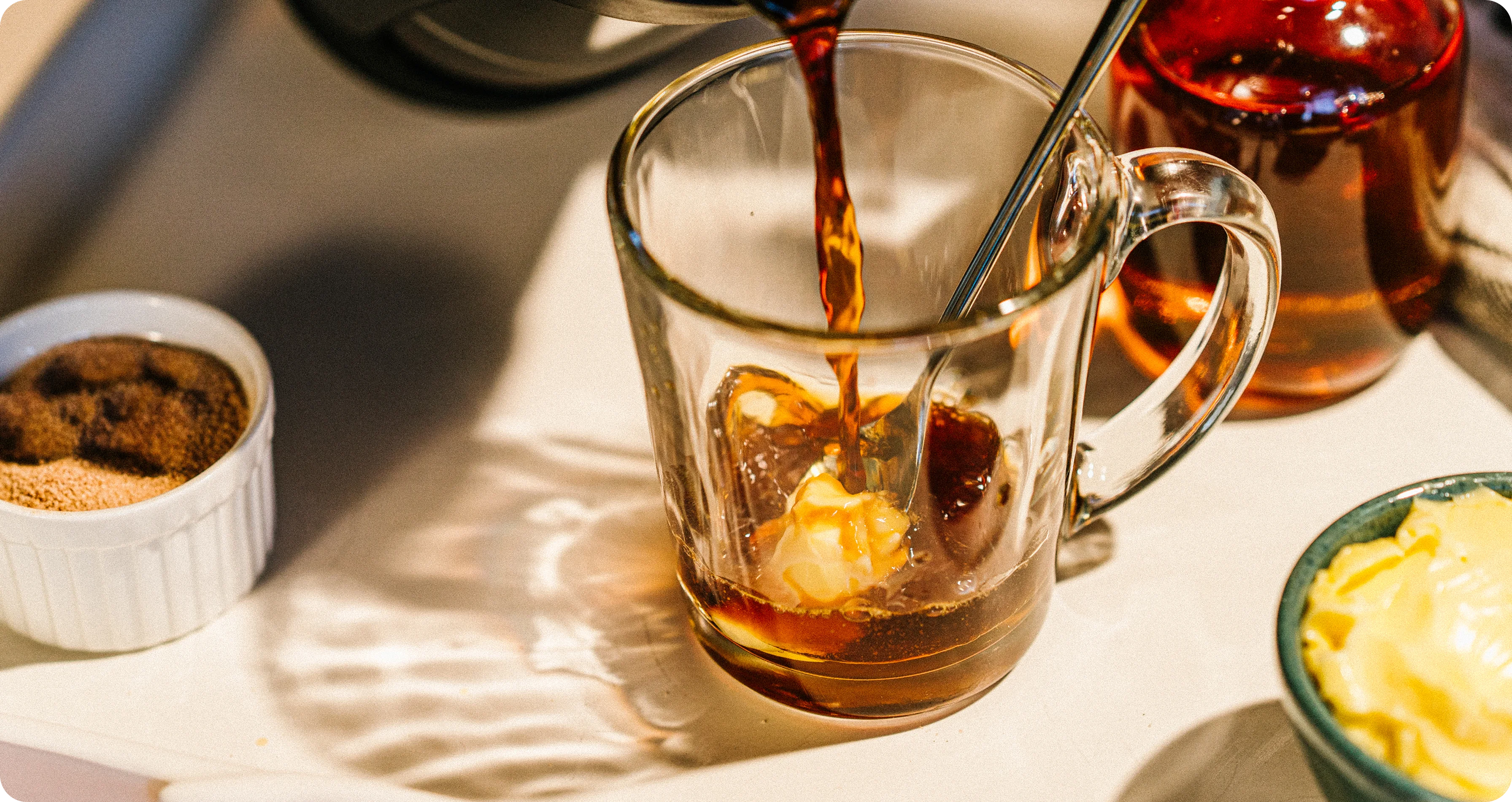 Glass mug with iced coffee being poured, surrounded by a dessert and a bowl of whipped cream.