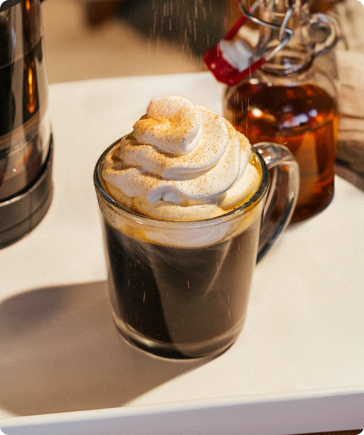 A glass mug filled with a dark coffee drink topped with a swirl of whipped cream and dusted with cinnamon, with syrup bottles in the background on a white surface.