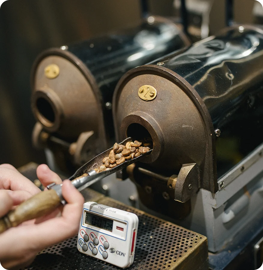 A close-up of a coffee roasting machine with roasted coffee beans being dispensed into a metal scoop held by a hand. A small digital timer is visible in the foreground.