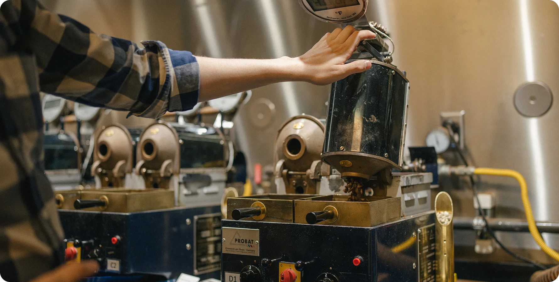A person in a blue and brown plaid shirt operates industrial coffee roasting machinery, with their hand resting on a dark metal hopper. The setting is a warm, industrial coffee roastery with stainless steel tanks and roasting machines in the background.