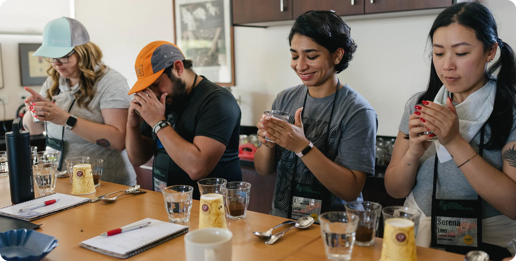 Four people sitting around a table in a cafe or workshop setting, each holding and examining a small smartphone. The table has glasses, notebooks, pens, and small containers, with a warm indoor background.