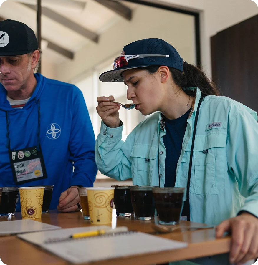A woman in a light blue jacket and navy cap tastes a dark beverage from a glass cup at a table, while a man in a blue shirt and black cap stands beside her. Paper cups, notebooks, and glasses are on the table in an indoor setting.