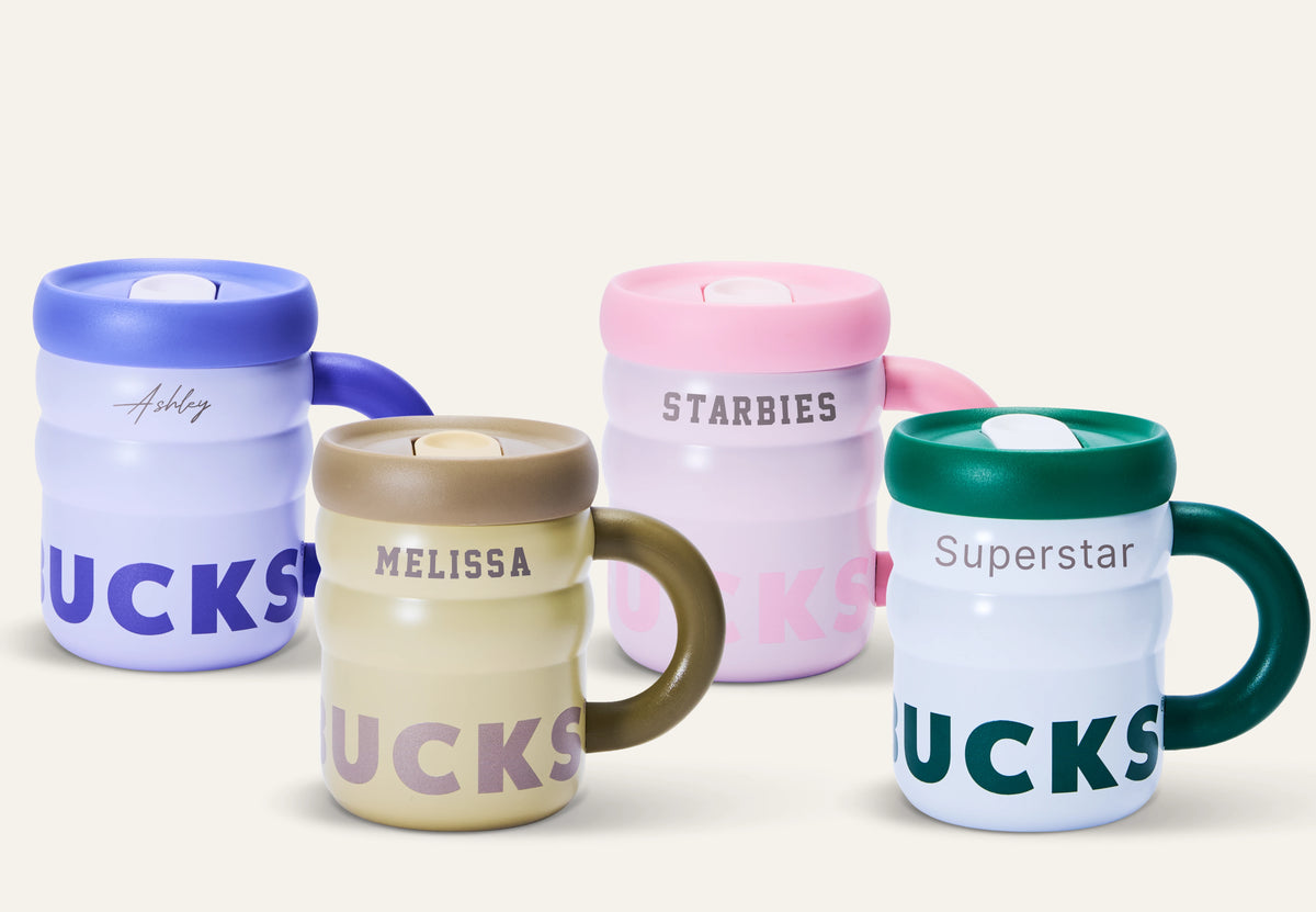 Four insulated travel mugs with lids are arranged on a light warm-gray studio background. The mugs are in pastel blue, cream, pink, and white, each with a different colored handle and lid.
