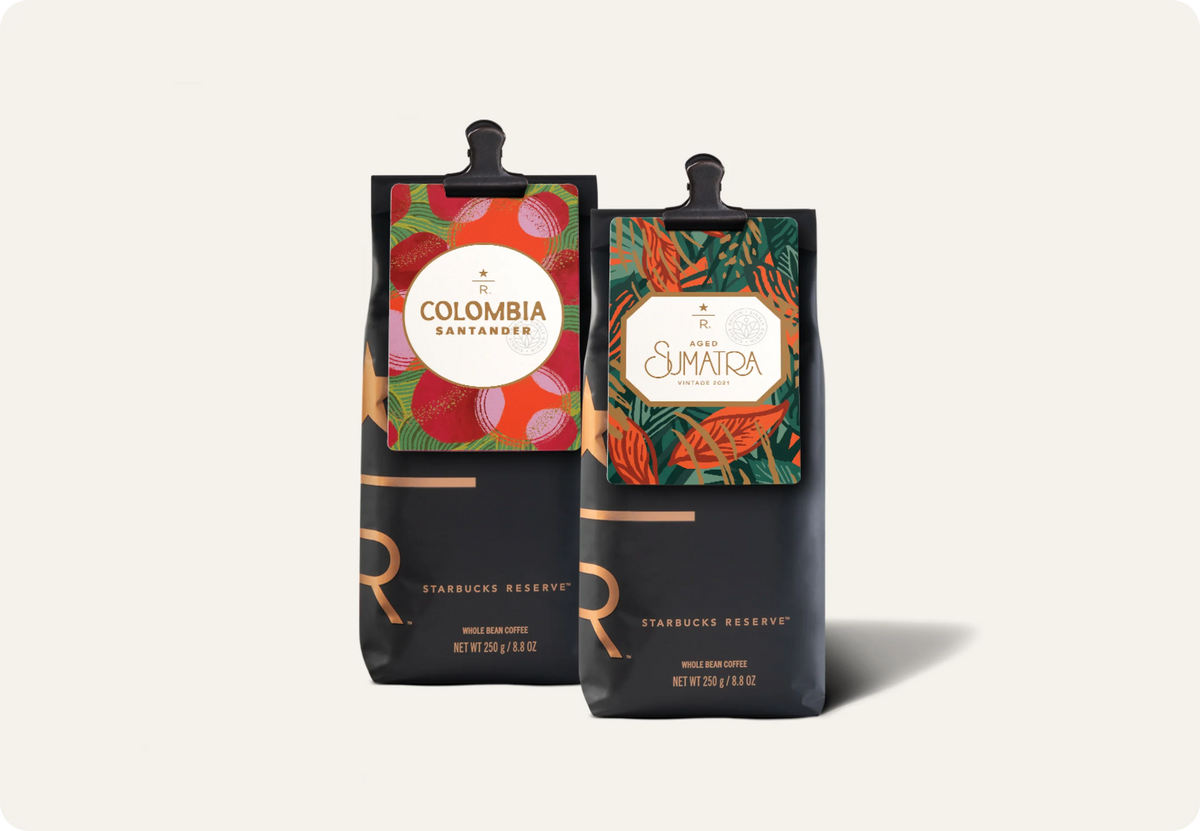 Two Starbucks Reserve coffee pouches standing upright on a light warm gray background. The left pouch is Colombia Santander with a red and orange botanical label, and the right pouch is Aged Sumatra Reserve with a green and orange botanical label.