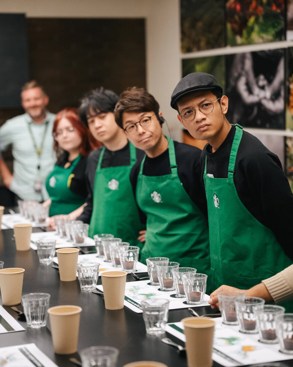 Group of Starbucks employees in green aprons at a training session with coffee cups and glasses on a table.